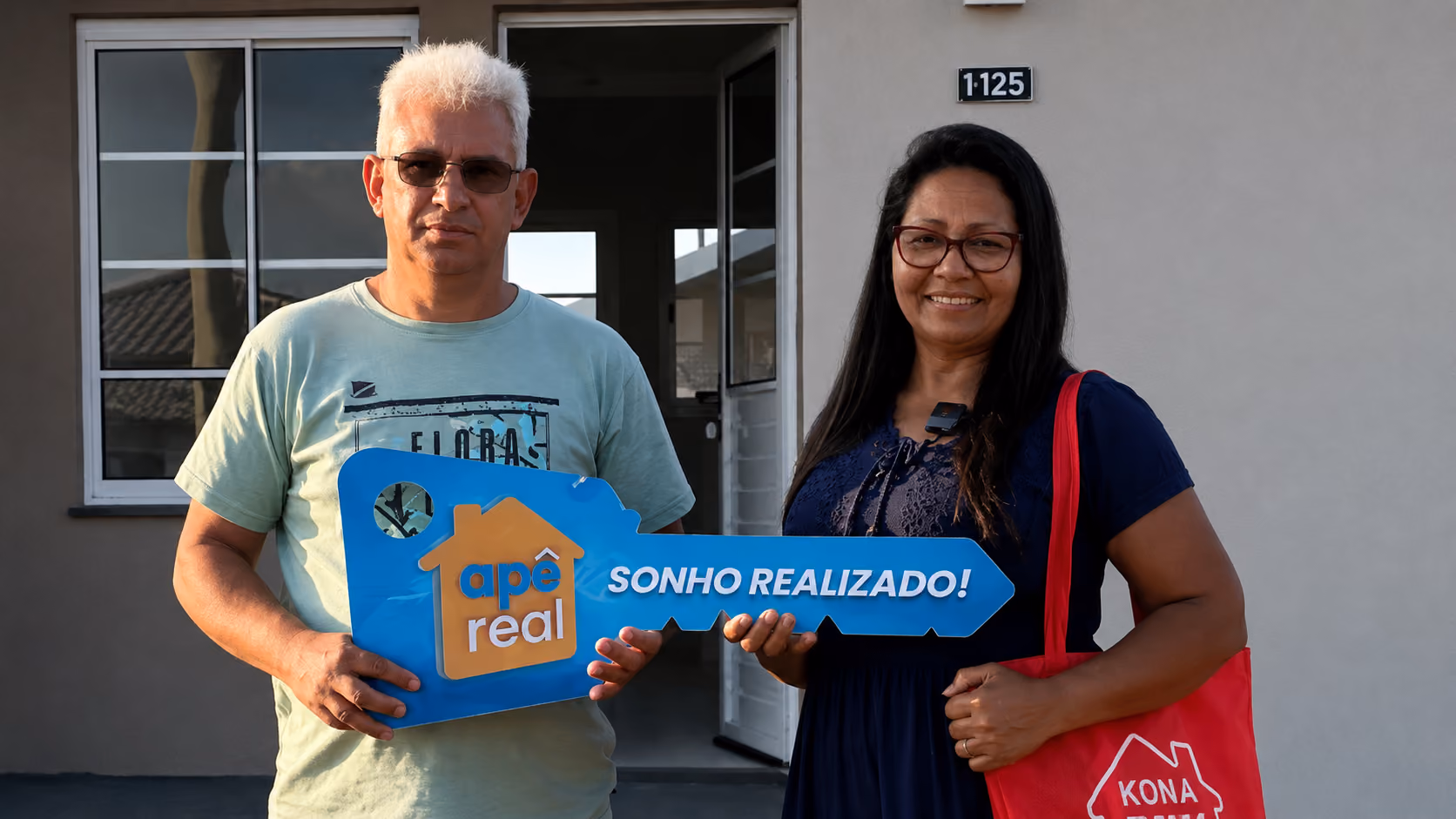 Casal sorridente segurando a placa-chave Apê Real na porta da casa própria que acabaram de conquistar