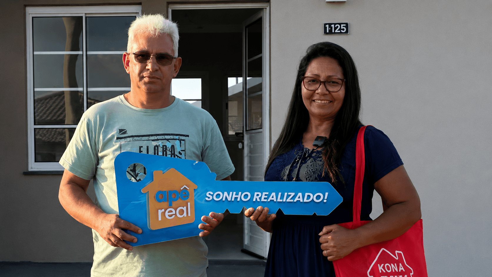 Casal sorridente segurando a placa-chave Apê Real na porta da casa própria que acabaram de conquistar